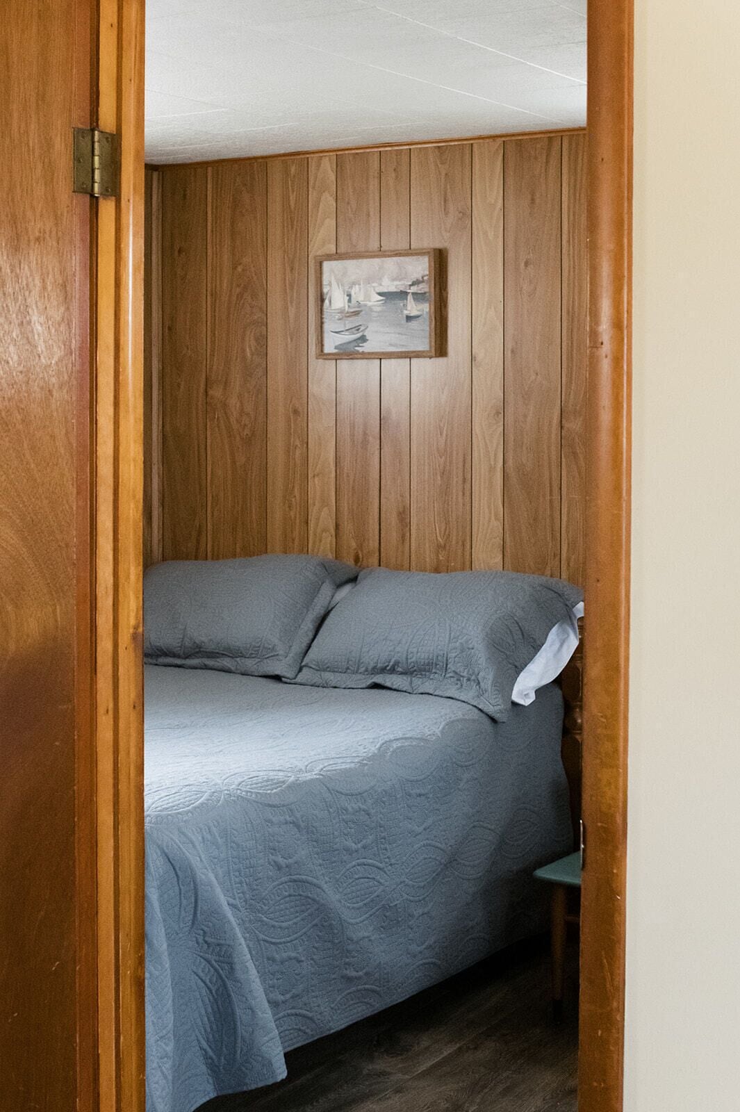 A bedroom with a wooden paneled wall, a bed with grey bedding, and a framed picture above the bed.