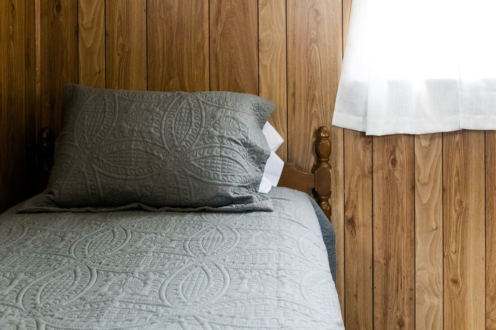 Un lit recouvert d’une couverture grise à motifs, avec un oreiller gris posé contre un mur en bois.
