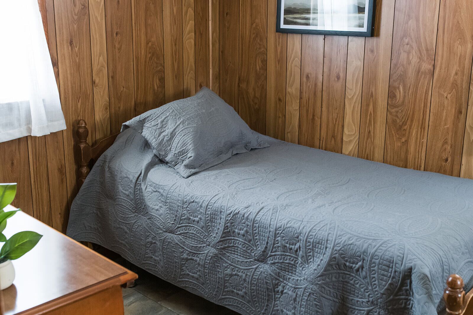 A single bed with a patterned bedspread and pillows, wooden panel walls, a framed picture on the wall, and a small table with a plant.