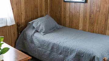 A single bed with a patterned bedspread and pillows, wooden panel walls, a framed picture on the wall, and a small table with a plant.