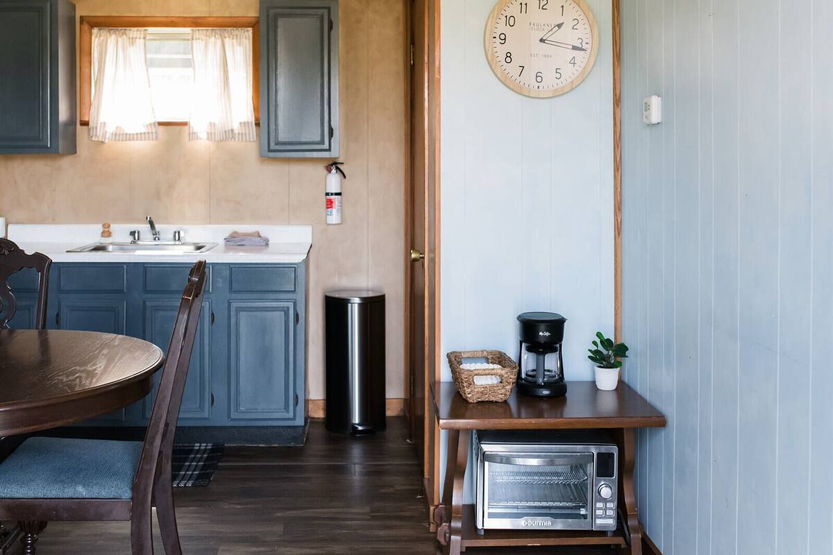 Een keuken met blauwe kastjes, een houten tafel, een koffiezetapparaat, een broodrooster en een wandklok.