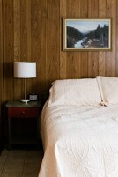 A bedroom with a wooden wall, a bed with a patterned bedspread, a bedside table with a lamp and a clock, and a framed landscape painting above the bed.