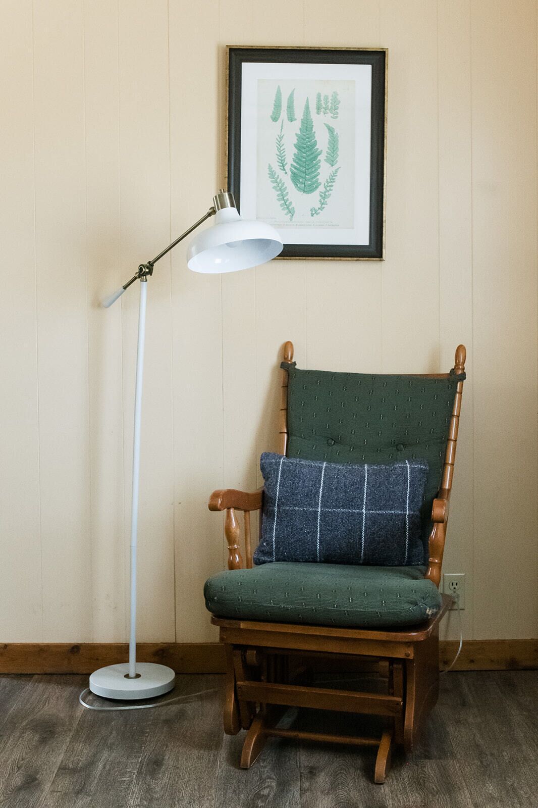 Une chaise en bois avec un coussin vert et un oreiller bleu, un lampadaire blanc et une estampe botanique encadrée accrochée au mur.
