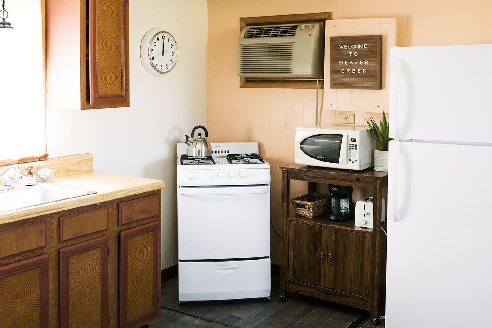 Una cocina equipada con estufa, microondas y refrigerador. Hay un reloj en la pared y un letrero de bienvenida.
