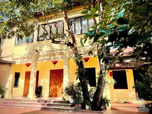 A yellow building with a staircase leading to the entrance, surrounded by potted plants and trees. - Lee Homestay (Hanoi)