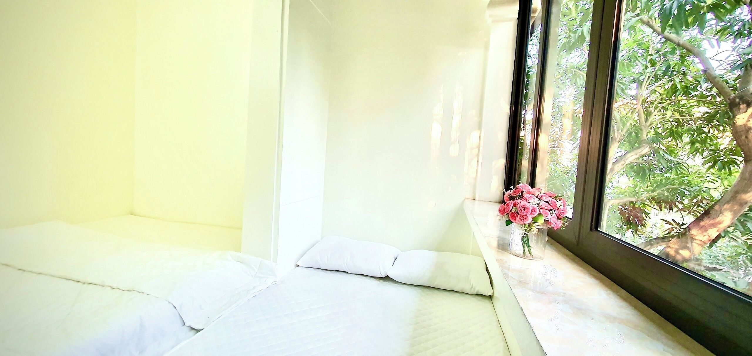 A room with a bed against a wall, a window with a view of trees, and a vase with flowers on the windowsill.