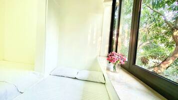 A room with a bed against a wall, a window with a view of trees, and a vase with flowers on the windowsill.