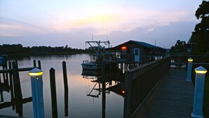 A wooden dock extending into calm water with a house on stilts and a boat docked nearby. - Boat Slip - Pool- Screened balcony- Water front. 30 DAY MINIMUM STAY. (Gulf Shores)