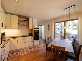 Eine moderne Küche mit integrierten Geräten, ein Essbereich mit Holztisch und Stühlen sowie große Fenster mit Jalousien.