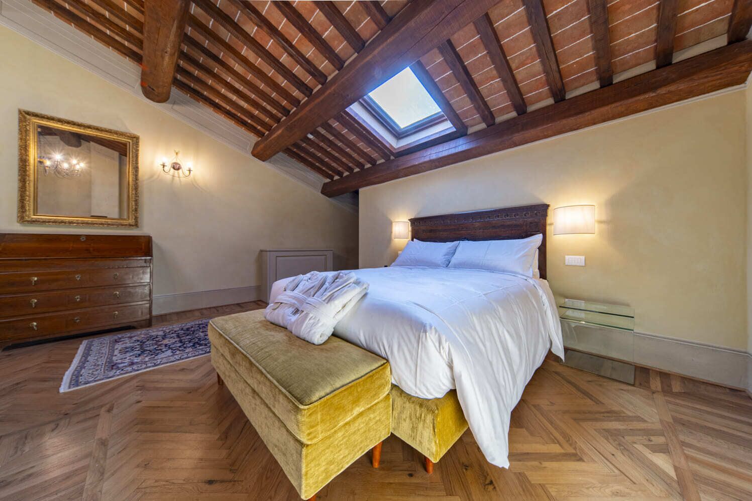 Quarto com teto de madeira, claraboia, cama com roupa de cama branca, cómoda de madeira e um banquinho.