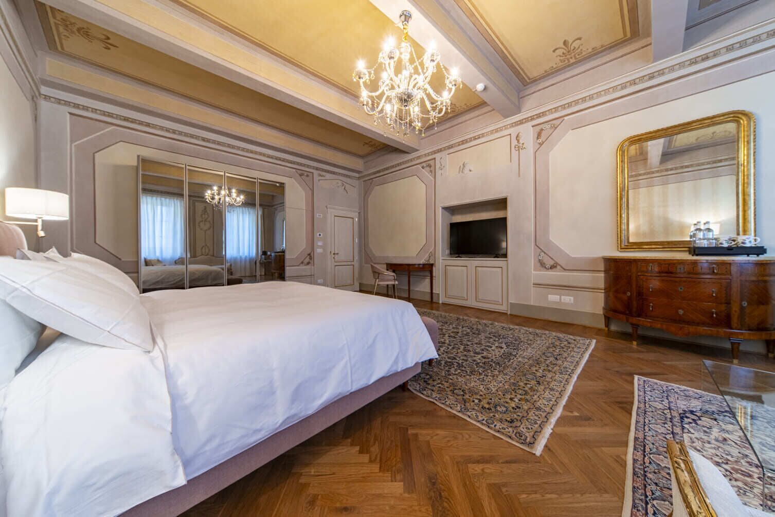 Een ruime slaapkamer met een groot bed, een kroonluchter, een spiegel, een televisie en een houten dressoir.