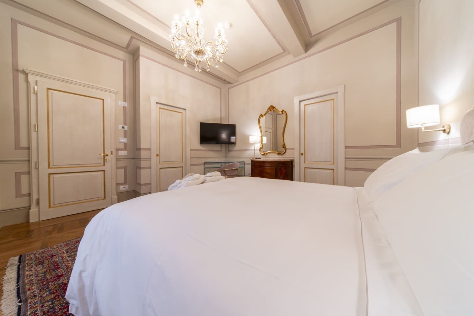 A large, neatly made bed in a hotel room with a television, a mirror, and a chandelier.