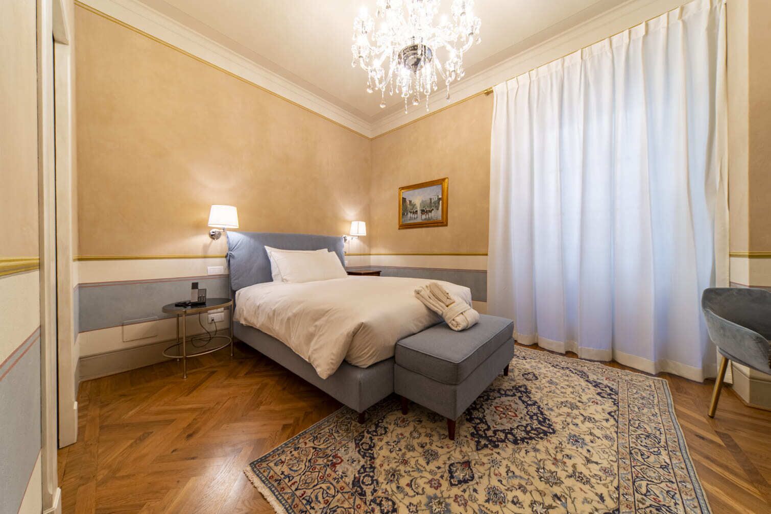 A hotel room with a bed, bedside lamps, a chandelier, a rug, and a chair.