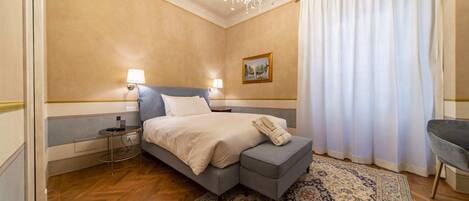 A hotel room with a bed, bedside lamps, a chandelier, a rug, and a chair.