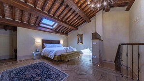 A bedroom with a large bed, a nightstand, a bedside table, a wooden floor, a ceiling with exposed beams, a skylight, a staircase, and a view of the outside.