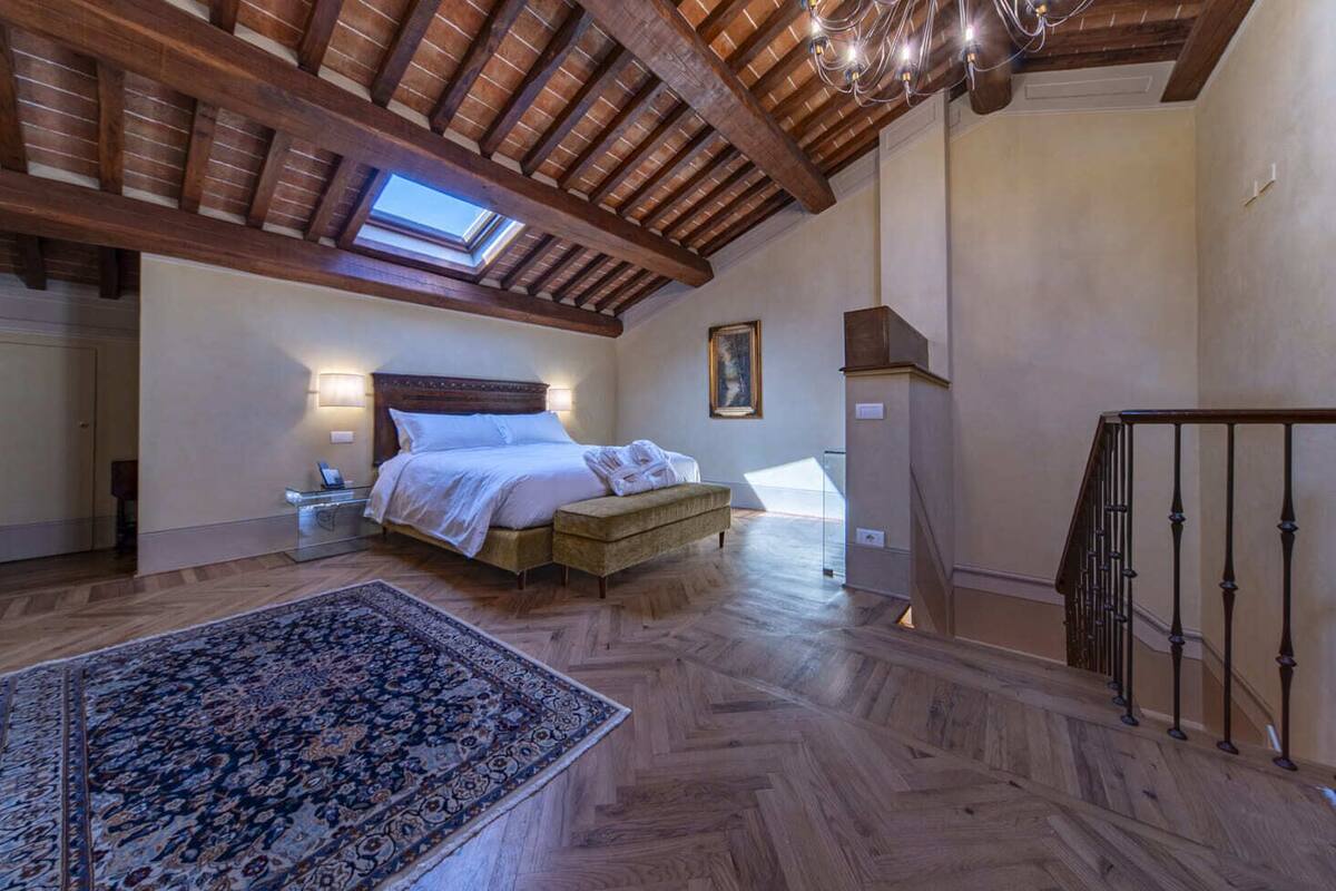 Un dormitorio con una cama grande, una mesita de noche, un tablero auxiliar, piso de madera, techo con vigas a la vista, tragaluz y vista al exterior.