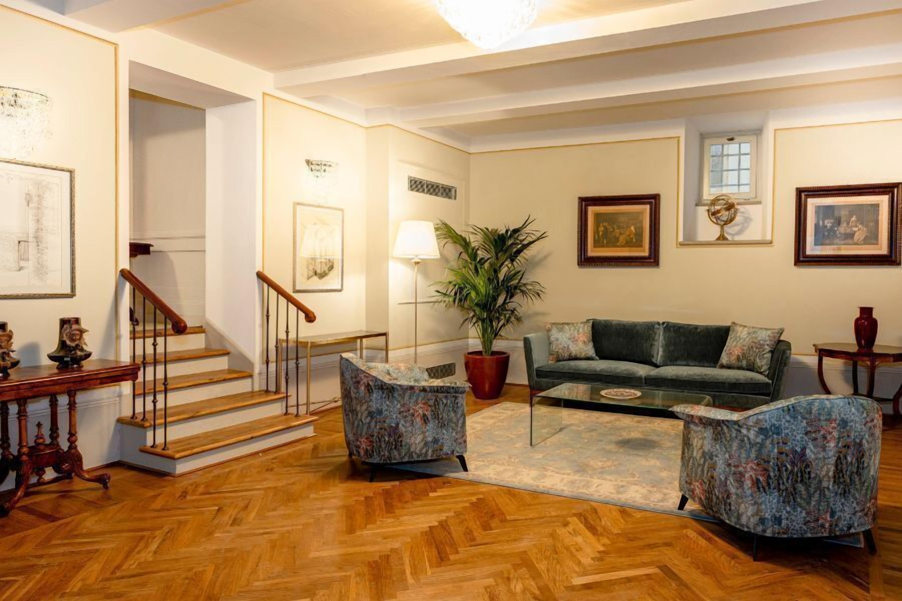 A spacious living room with a herringbone floor, a green sofa, two patterned armchairs, a coffee table, and a staircase with wooden railings.
