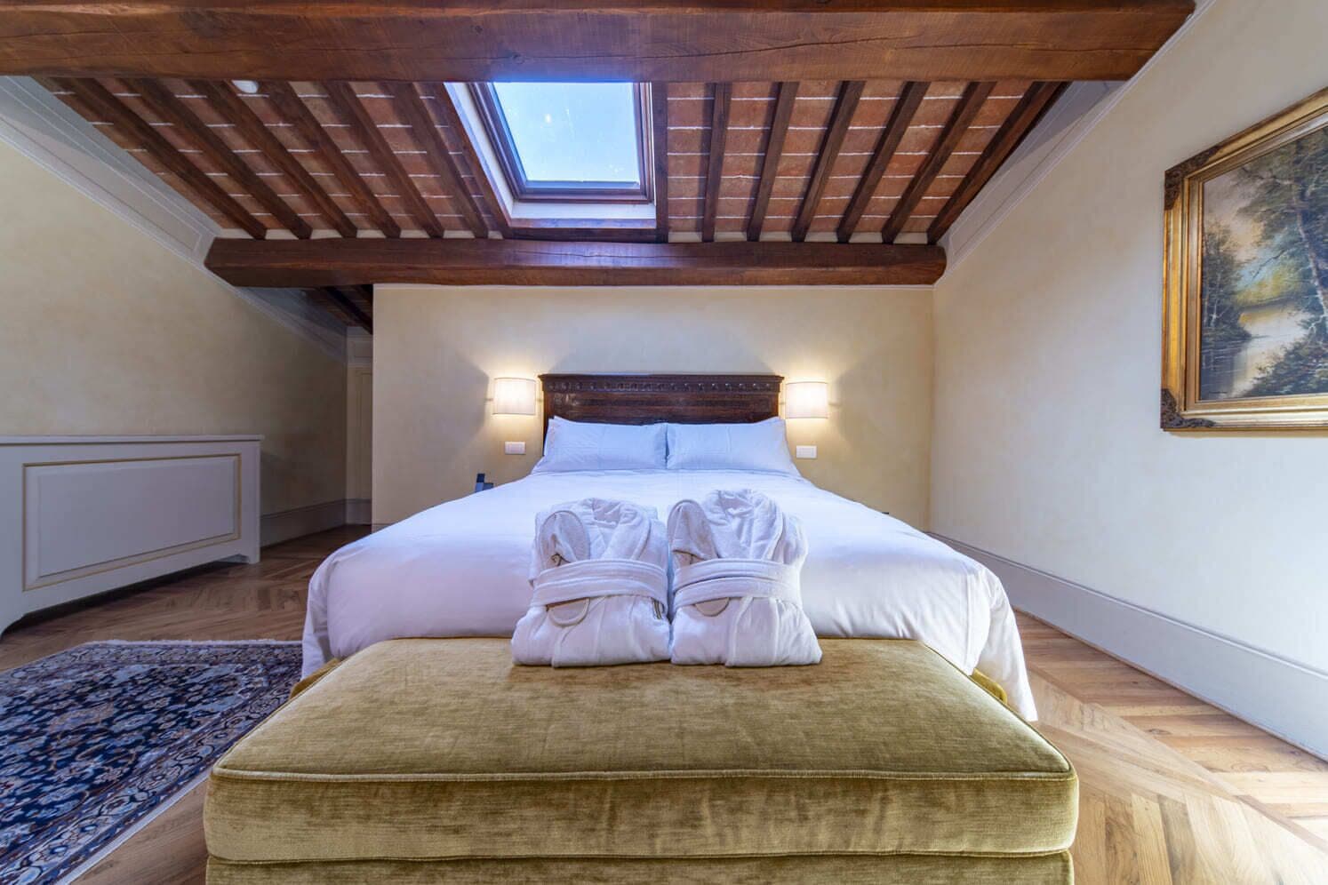 A bedroom with a skylight, a bed with white linens, a pair of towels on an ottoman, and a painting on the wall.