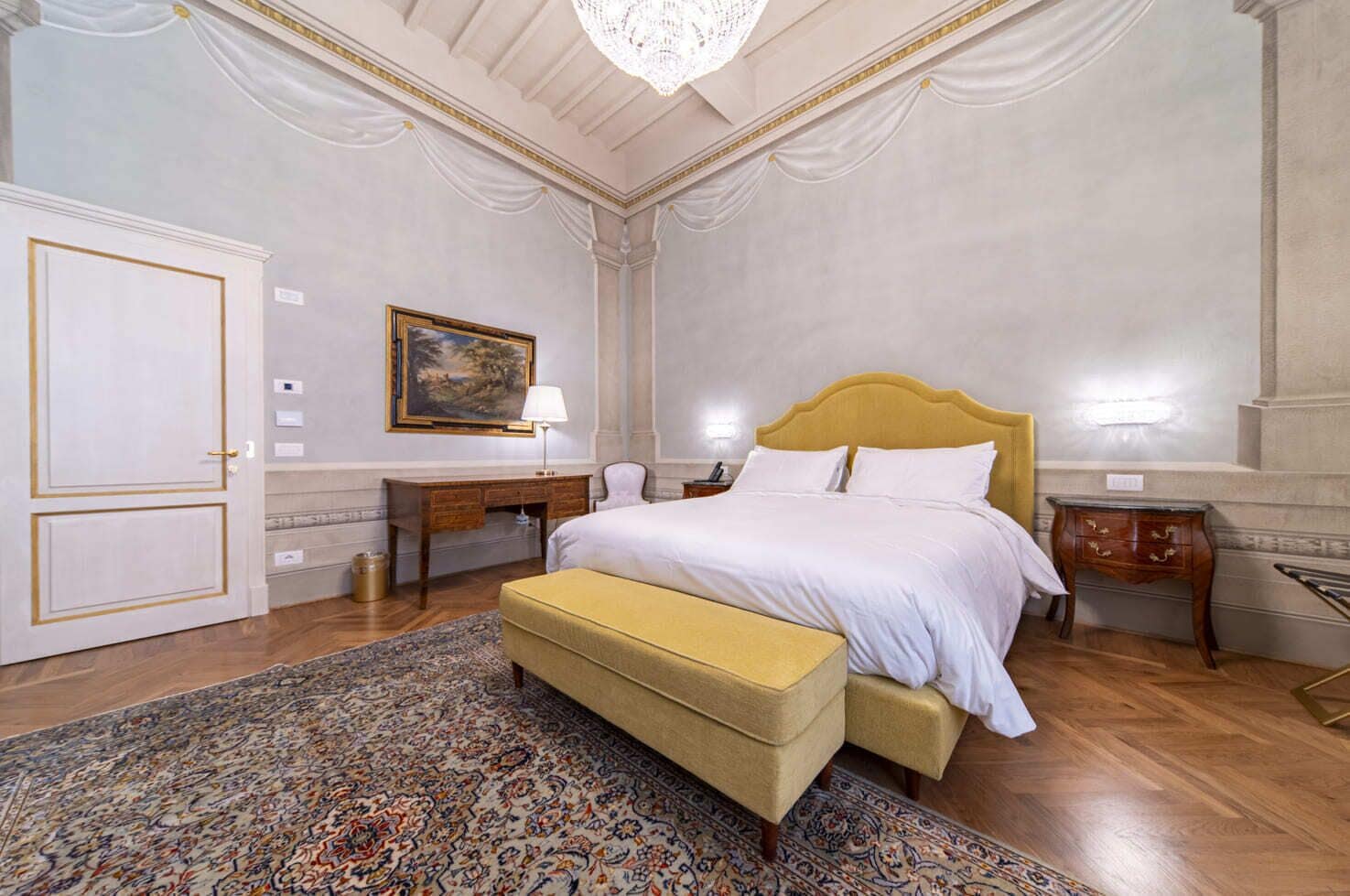 A bedroom with a large bed, a desk, a chair, a bench, and a patterned rug.