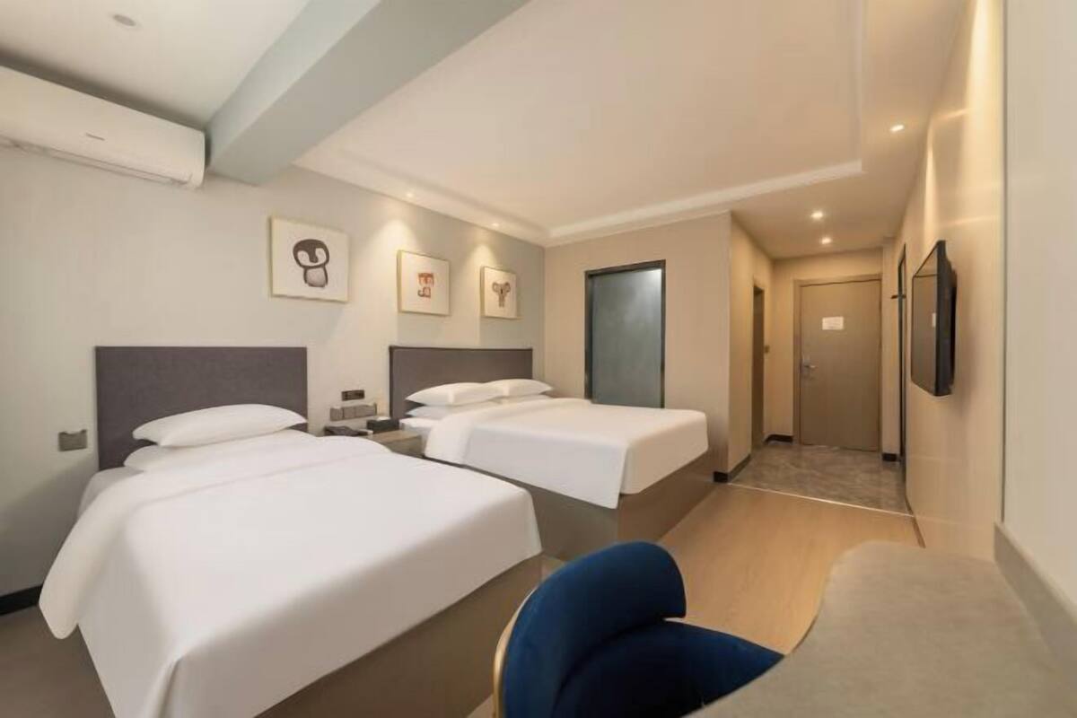 Een hotelkamer met twee bedden, een bureau en een stoel. Er hangen prenten aan de muur, en er is een deur en een raam.
