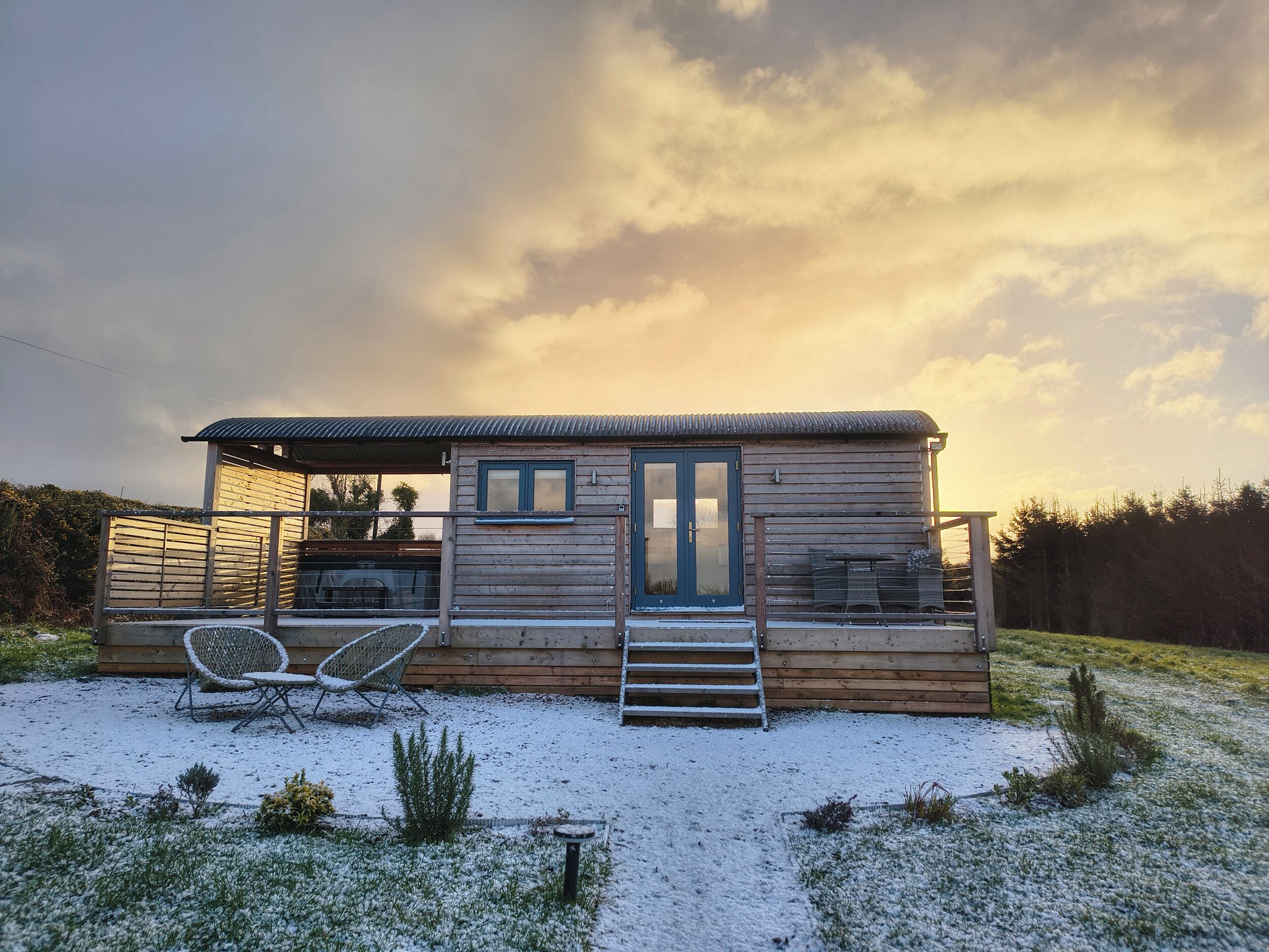 A wooden cabin with a porch, a set of stairs leading to the entrance, and two chairs outside.