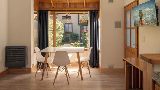 A dining area with a table and chairs, a view of a garden, and a painting on the wall. - Departamento en el centro para 4 pax (San Martín de los Andes)