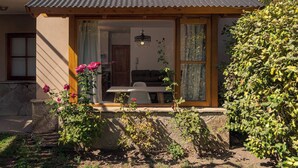 A cozy room with a wooden table and chairs, a hanging light fixture, and a view of a garden with pink flowers. - Departamento en el centro para 4 pax (San Martín de los Andes)