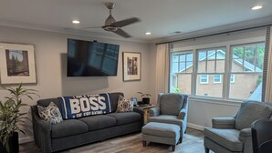 A living room with a gray sofa, two armchairs, a coffee table, and a ceiling fan. There are framed pictures on the walls and a large window with a view of a brick building. - Charming studio in lovely Charlotte with WiFi, AC (Charlotte)