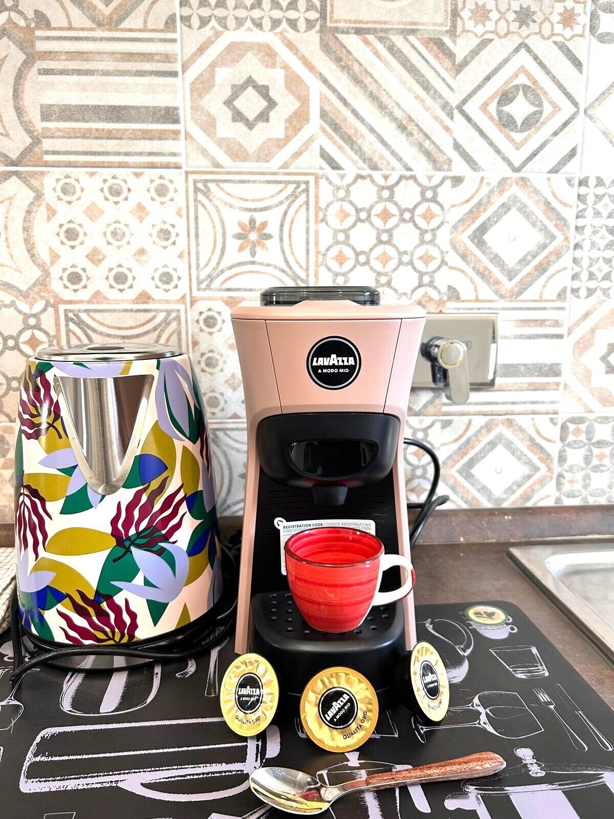 A coffee maker with a registration code, a colorful kettle, and a chalkboard with utensils drawn on it.