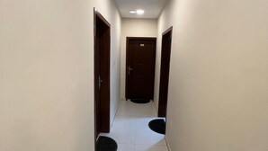 Un couloir étroit et bien éclairé, avec un sol carrelé de carreaux blancs et des portes en bois de chaque côté.
