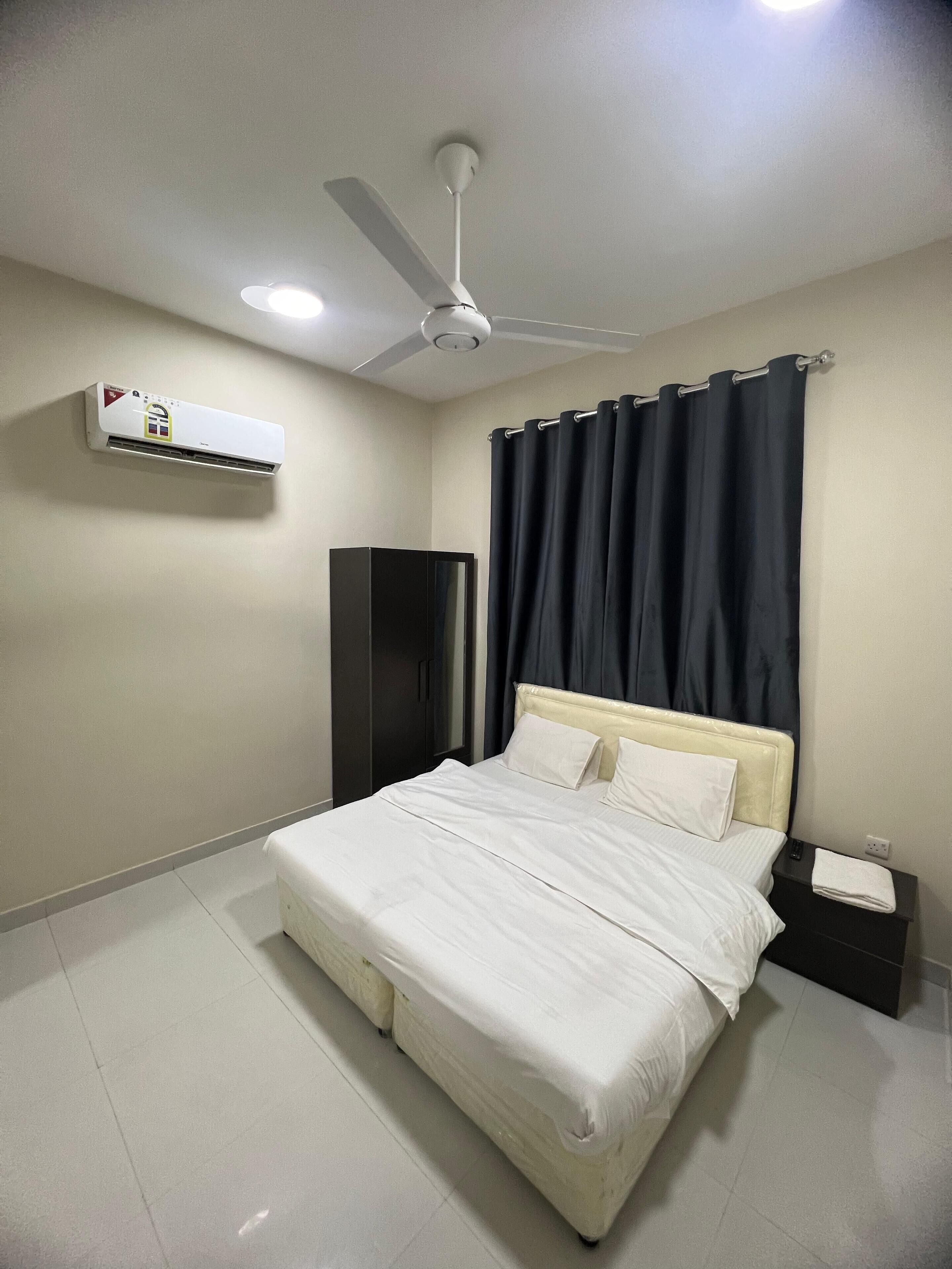 Une chambre à coucher comprenant un lit, un ventilateur de plafond, un appareil de climatisation, une table de chevet et une armoire de couleur sombre.