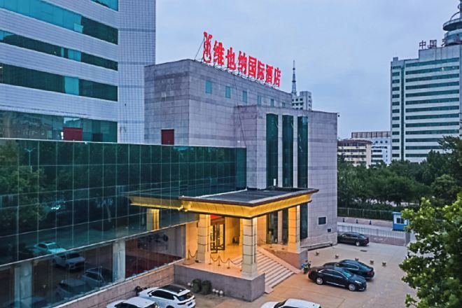 A modern building with a glass facade, a prominent entrance with steps, and a parking area with cars.
