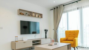 A modern living room with a flat-screen TV, a yellow armchair, a white coffee table, and a minimalist console table with decorative items. - Snug three-bedroom Unit (Accra)