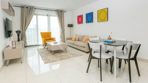 A modern living room with a sofa, a yellow armchair, a coffee table, and a dining table with chairs. There is a flat-screen TV mounted on the wall, a floor lamp, and three framed artworks on the wall. - Snug three-bedroom Unit (Accra)