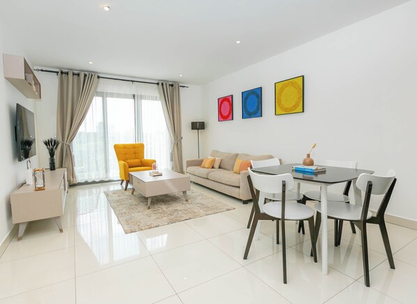 Uma sala de estar moderna com um sofá, uma poltrona amarela, uma mesa de centro e uma mesa de jantar com cadeiras. Há uma televisão de tela plana na parede, um abajur e três obras de arte emolduradas.