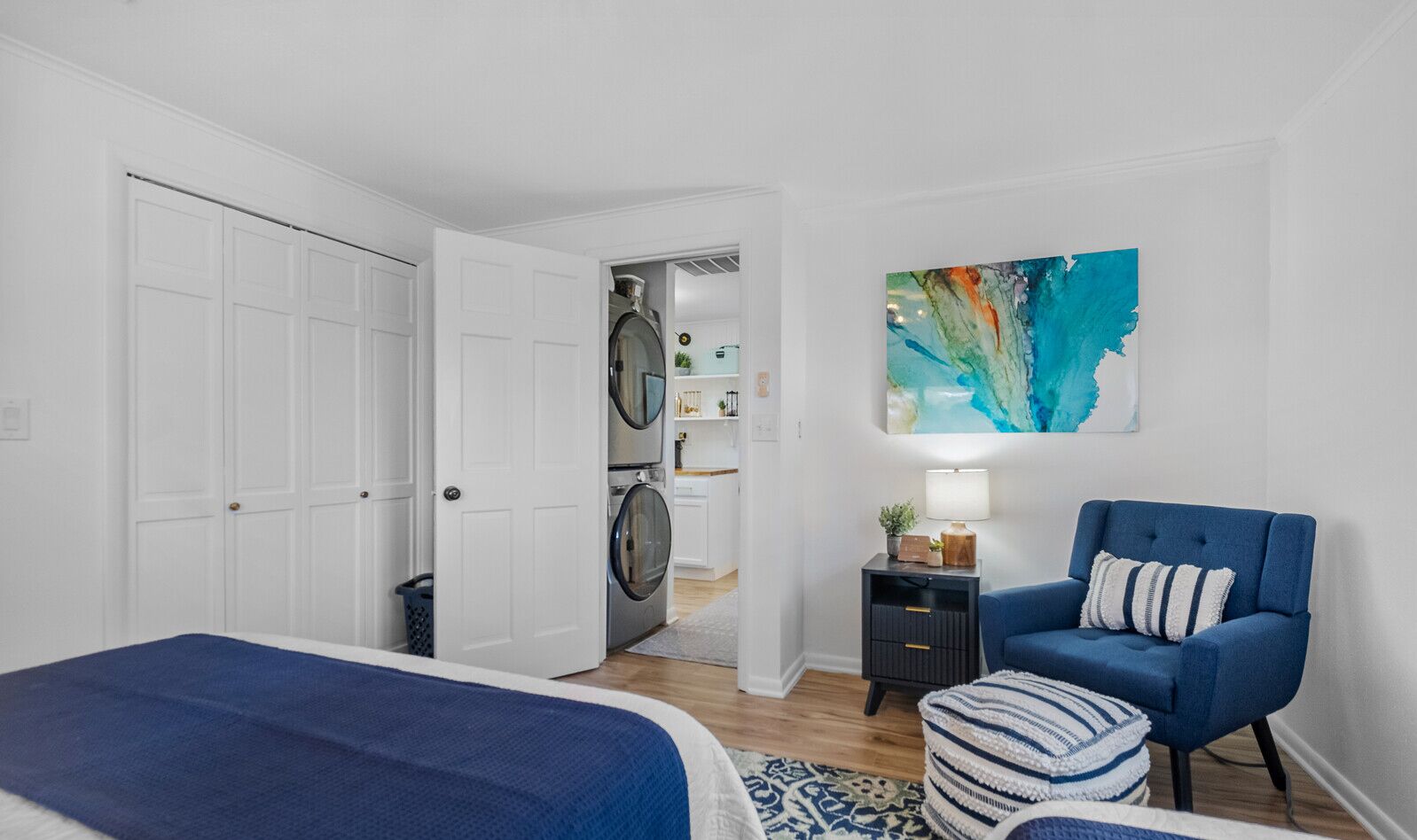 A bedroom with a bed, a chair, a nightstand, a painting, and a laundry area.