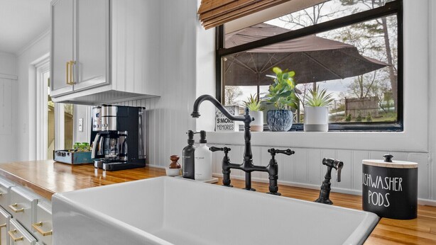A modern kitchen with a large white sink, black faucets, and a coffee machine. There are potted plants on the windowsill and a sign that says "AMAZING MEMORIES."