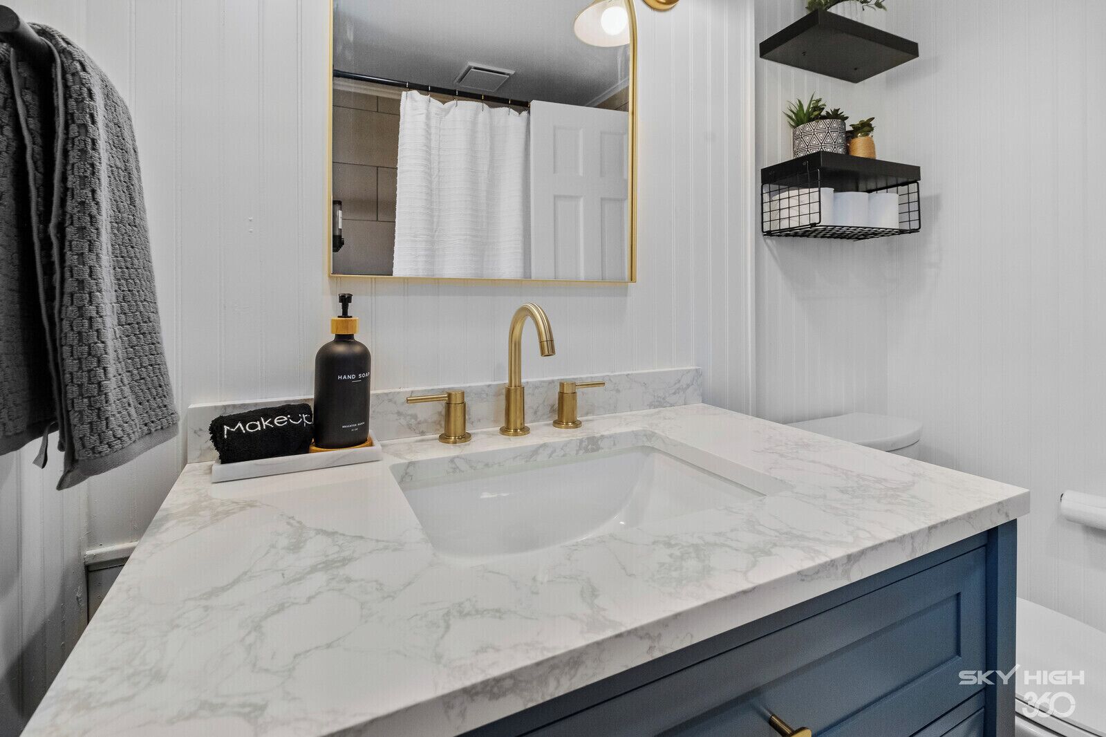 Une salle de bain avec un lavabo en marbre blanc, des robinets dorés, un distributeur de savon noir, un porte-serviettes et un miroir.