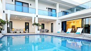 Un espace piscine intérieur moderne, doté d’une piscine à l’eau d’un bleu limpide, de chaises longues et d’un coin salon avec des tables et des chaises.
