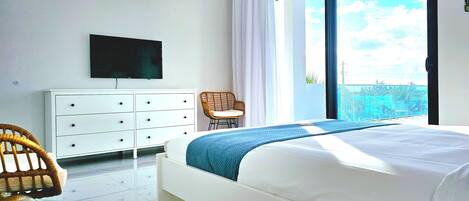 A bedroom with a bed, a TV, a dresser, a chair, and a large window with a view.
