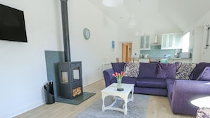 A modern living room with a fireplace, a sofa, a coffee table, and a kitchen in the background. - Cornel Clud (Cemaes Bay)