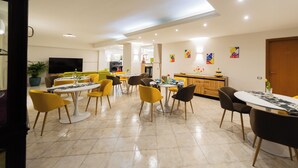 Een ruime eetruimte met meerdere tafels en stoelen, een keuken met een houten aanrecht, en een tv aan de muur.