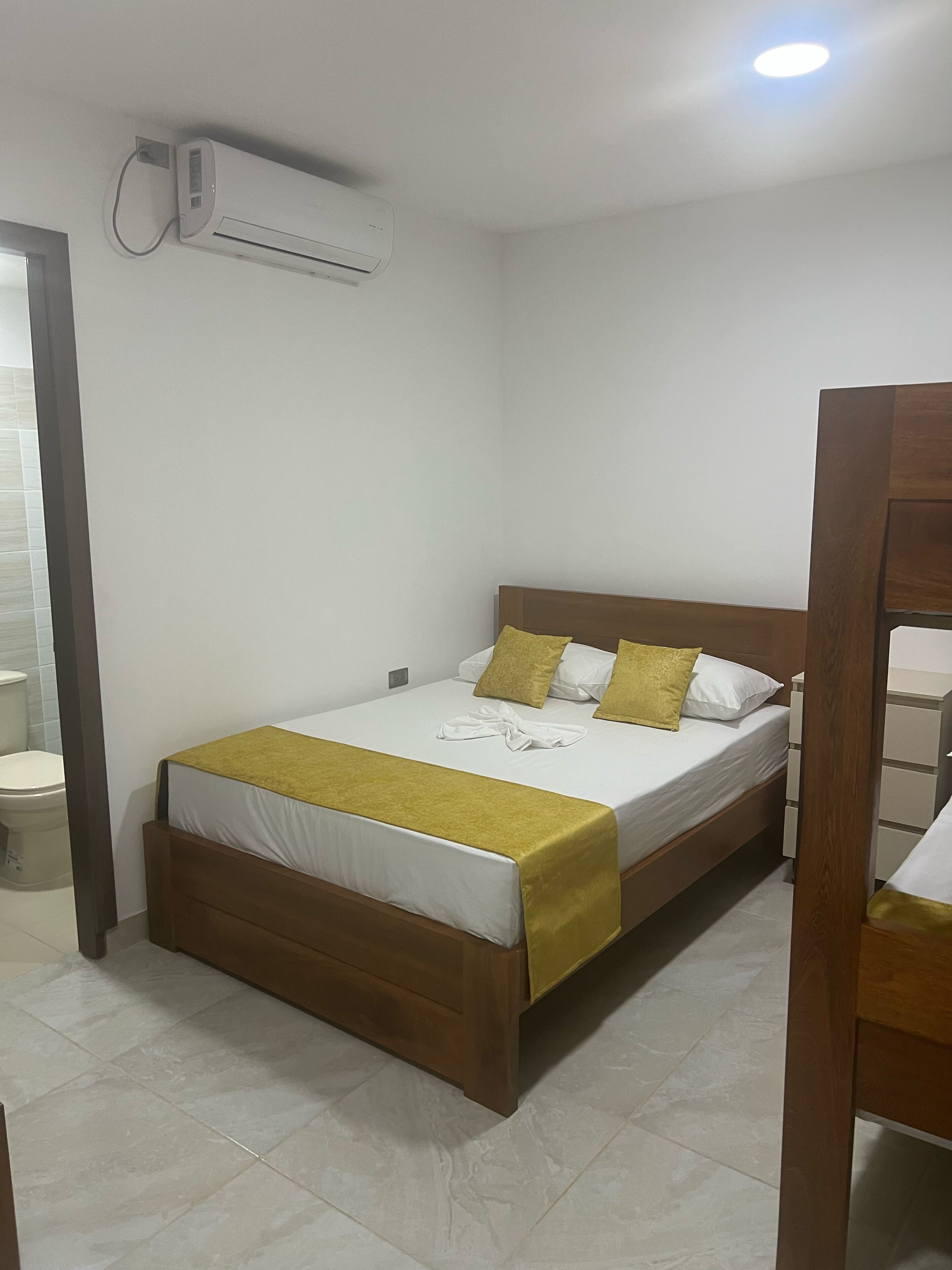 Habitación individual con una cama y cabecera de madera, ropa de cama blanca y una manta amarilla. Un equipo de aire acondicionado está montado en la pared sobre la cama.