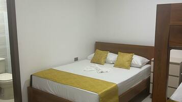 Habitación individual con una cama y cabecera de madera, ropa de cama blanca y una manta amarilla. Un equipo de aire acondicionado está montado en la pared sobre la cama.