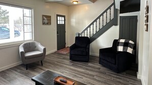 A living room with a grey armchair, a black armchair, and a coffee table with a remote control. A staircase with a black railing leads to another room. - Charming 3-bedroom house in Trout Lake close to trails & lakes. (Trout Lake)