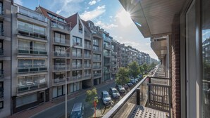 Un immeuble d’habitation doté de nombreux balcons, une rue bordée de voitures garées et un ciel dégagé.