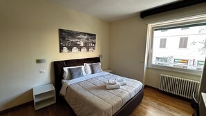 A bedroom with a bed, a nightstand, a window with a view of buildings, and a wall-mounted picture. - rienzo vatican suites  (Rome)