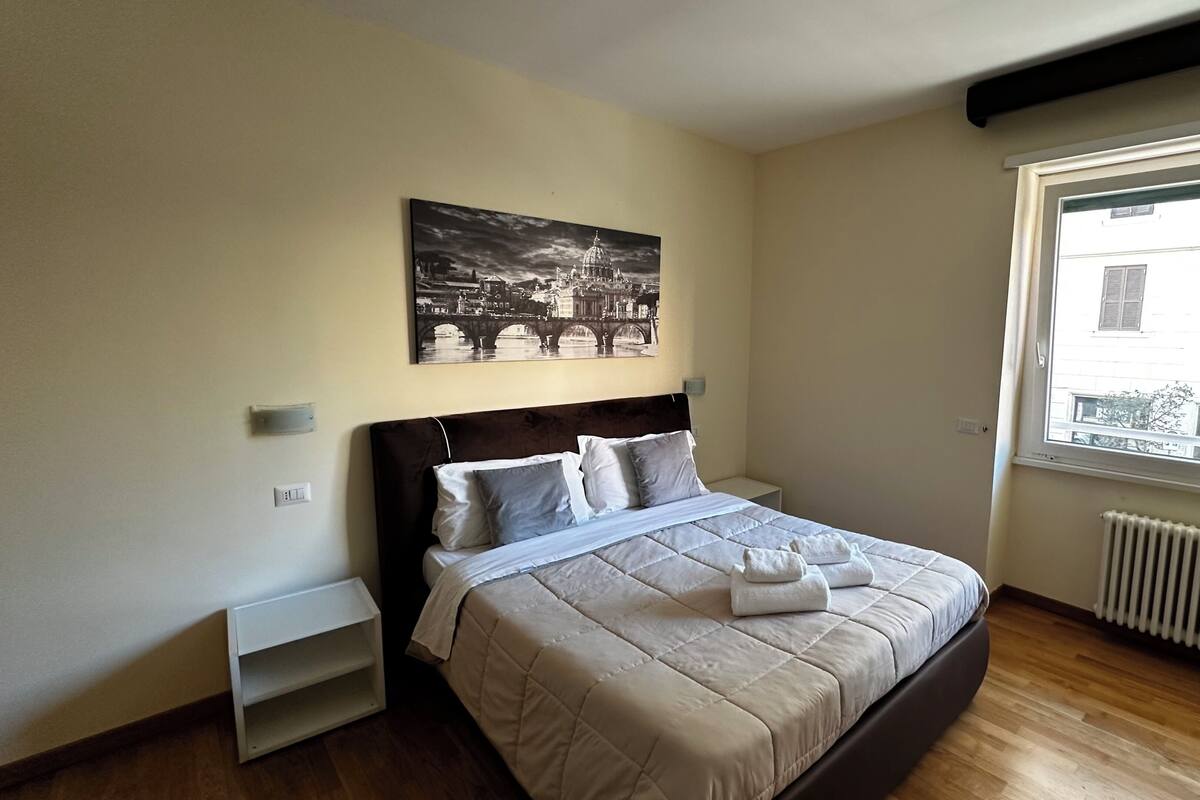 A bedroom with a bed, a nightstand, a window with a view of buildings, and a wall-mounted picture.