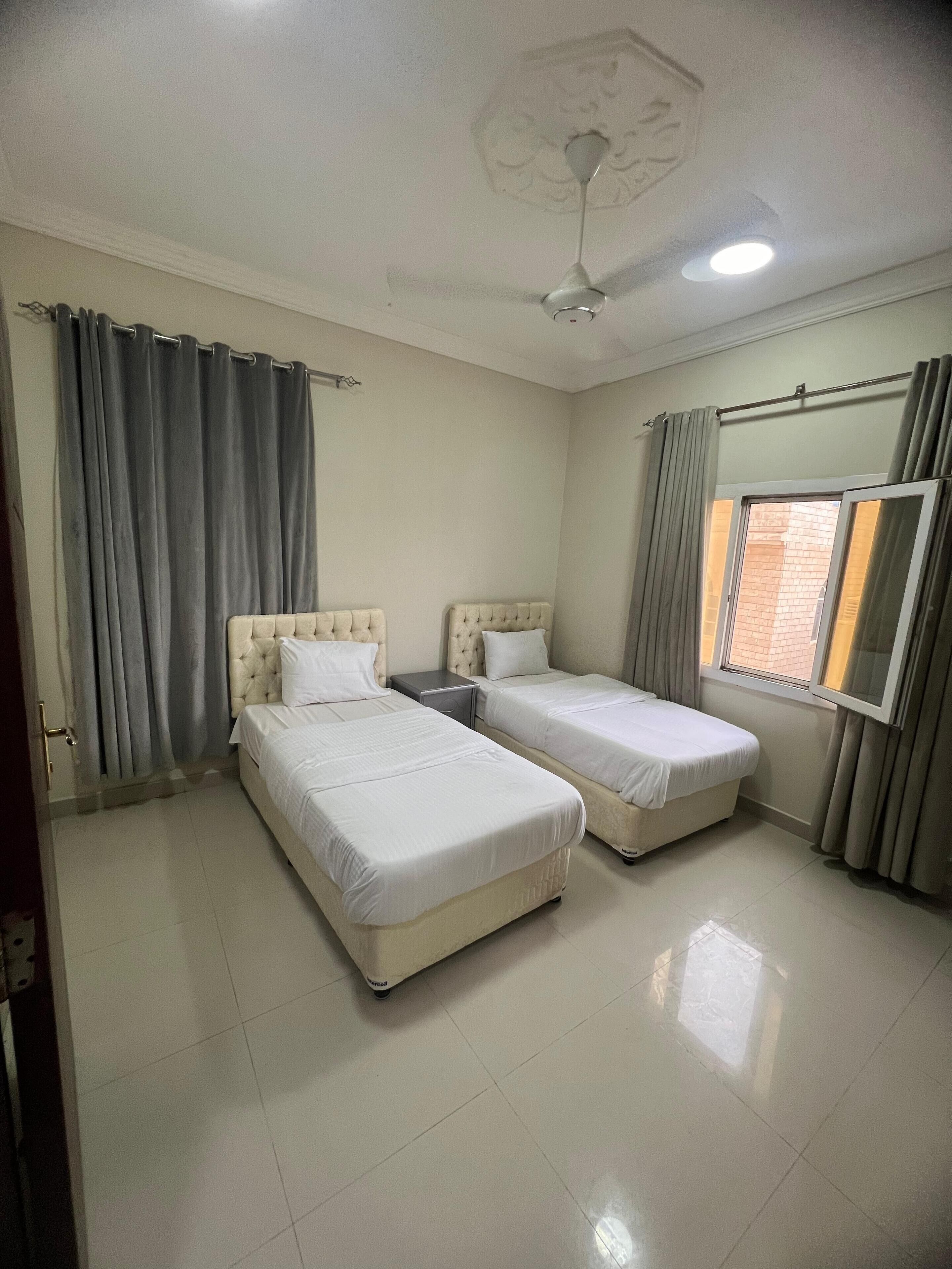 A room with two beds, a ceiling fan, a window with curtains, and a door.