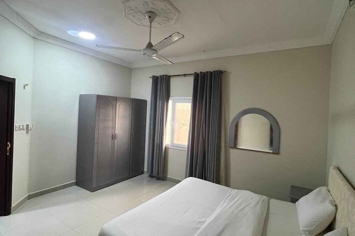 Une chambre à coucher comprenant un lit, une armoire, un ventilateur de plafond, une fenêtre avec des rideaux et un miroir au mur.